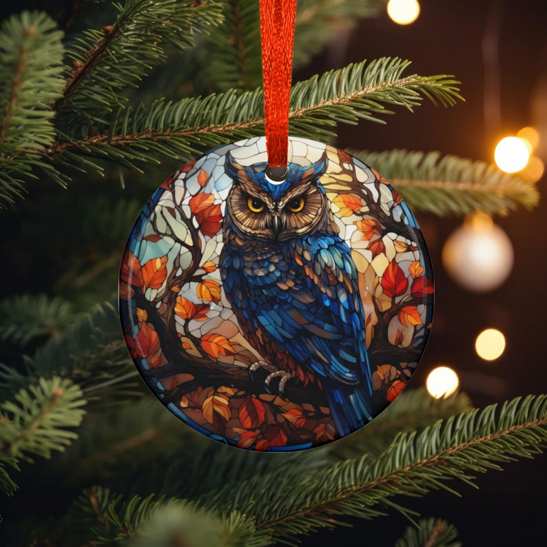 May include: A round Christmas ornament featuring a detailed illustration of a blue owl perched on a branch with autumn leaves. The ornament has a red ribbon for hanging and is set against a backdrop of a Christmas tree.