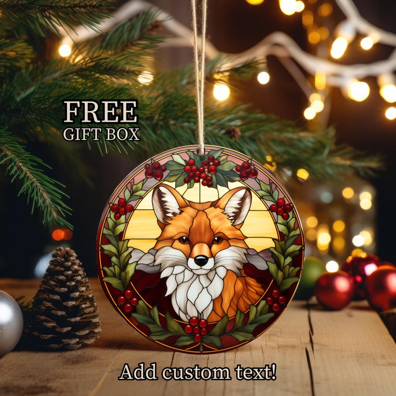 May include: A round stained glass Christmas ornament featuring a red fox with a holly wreath and red berries. The ornament is hanging from a string and has a white background. "FREE GIFT BOX" is written in black text at the top of the image. "Add custom text!" is written in black text at the bottom of the image.