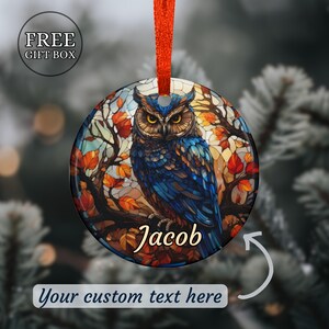 May include: Round Christmas ornament featuring a blue owl with yellow eyes, perched on a branch with autumn leaves. The ornament has the name "Jacob" and the text "Your custom text here". It includes a red ribbon for hanging.