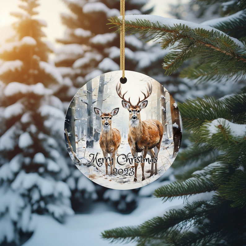 May include: A round Christmas ornament with a winter forest scene featuring two deer. The deer are brown and white with antlers. The text "Merry Christmas 2023" is printed on the ornament.