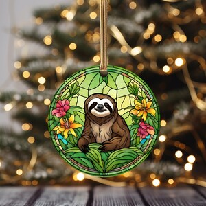 May include: A round glass ornament with a stained glass design featuring a brown sloth sitting in a green jungle with yellow and pink flowers.