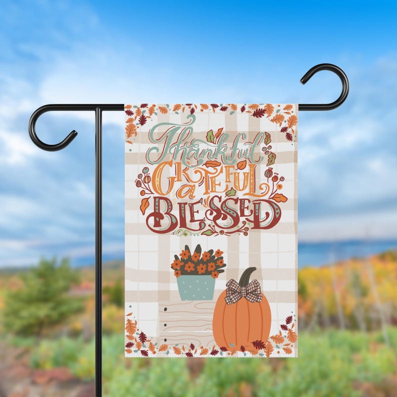 May include: A garden flag with the words "Thankful, Grateful, Blessed" in a decorative font. The flag features a pumpkin, flowers, and fall leaves on a plaid background. The flag is hanging on a black metal stand.