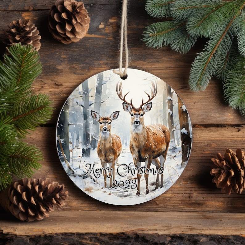May include: A round Christmas ornament featuring a snowy forest scene with two deer. The deer are brown with white spots and are standing in the snow. The text "Merry Christmas 2023" is printed on the ornament.