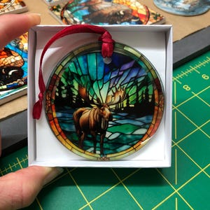 K&ouml;nnte beinhalten: Ein runder Glasanh&auml;nger mit einem Buntglasdesign eines Elchs, der in einem Wald steht. Der Anh&auml;nger befindet sich in einer wei&szlig;en Schachtel mit einem roten Band.