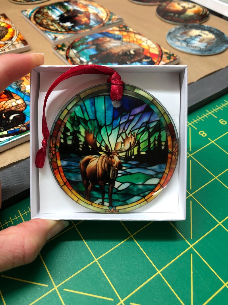 K&ouml;nnte beinhalten: Ein runder Glasanh&auml;nger mit einem Buntglasdesign eines Elchs, der in einem Wald steht. Der Anh&auml;nger befindet sich in einer wei&szlig;en Schachtel mit einem roten Band.