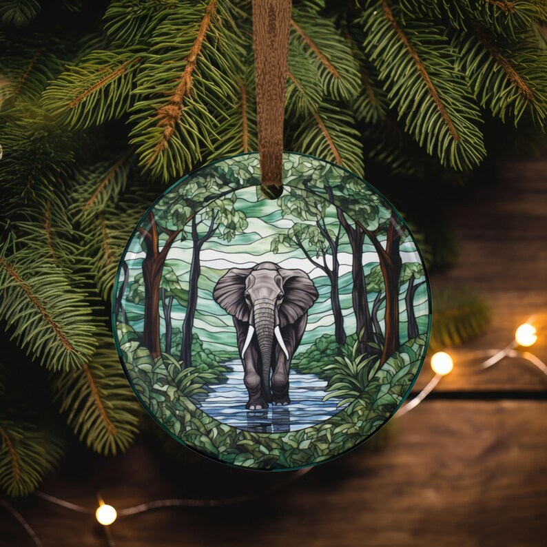 May include: A round Christmas ornament with a detailed illustration of an elephant walking in a river surrounded by trees. The ornament has a brown ribbon for hanging and is set against a backdrop of evergreen branches.
