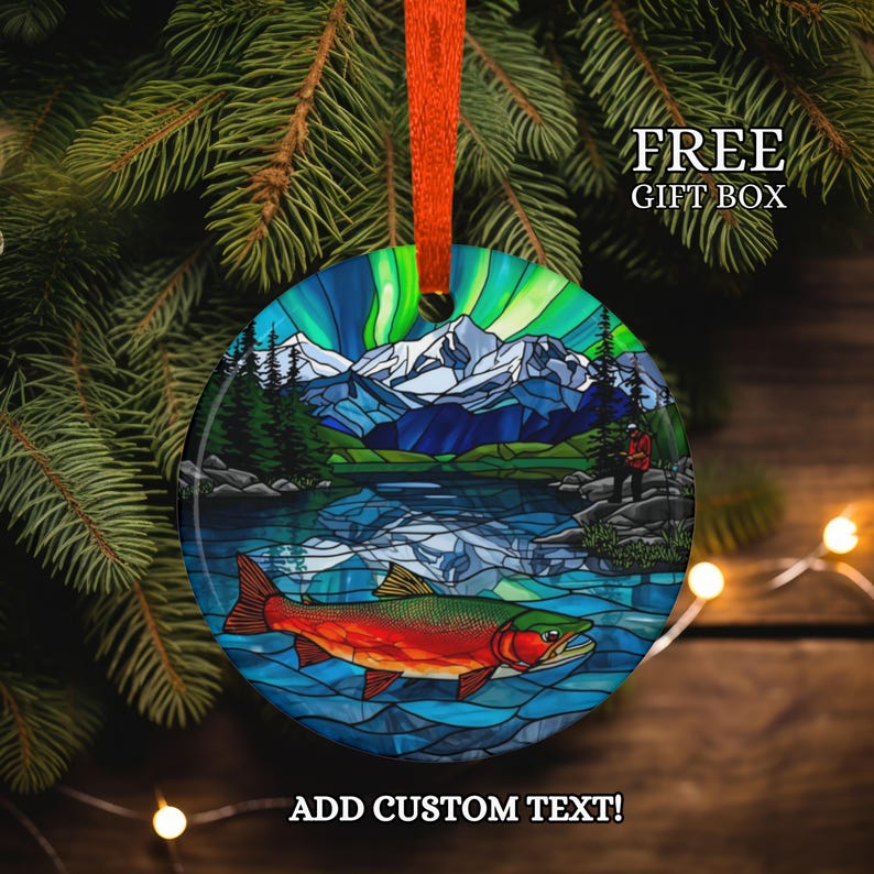 May include: Round Christmas ornament with a colorful stained-glass design featuring a lake scene with a red and green fish, mountains, and the Northern Lights. The ornament has an orange ribbon for hanging and the text "FREE GIFT BOX" and "ADD CUSTOM TEXT!"