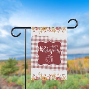 K&ouml;nnte beinhalten: Eine Thanksgiving-Gartenflagge mit einem rot-wei&szlig; karierten Muster und einem weinroten zentralen Design. Das Design zeigt die Worte "Happy Thanksgiving" in Schreibschrift und eine wei&szlig;e K&uuml;rbisillustration. Florale Akzente in Herbstfarben schm&uuml;cken die Ober- und Unterseite.