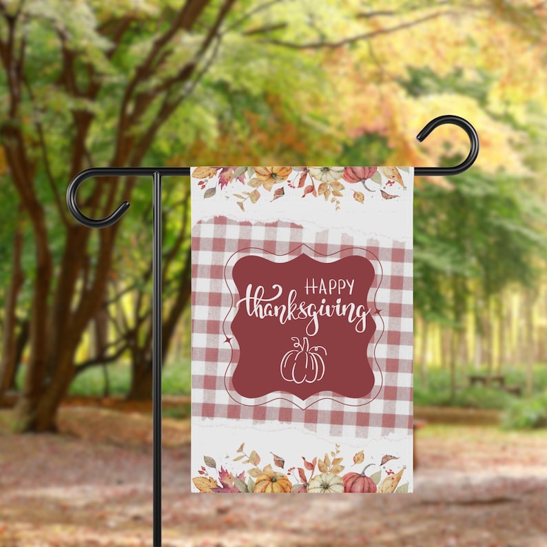 K&ouml;nnte beinhalten: Eine Thanksgiving-Gartenflagge mit einem rot-wei&szlig; karierten Muster und einem weinroten zentralen Design. Das Design zeigt die Worte "Happy Thanksgiving" und eine K&uuml;rbisillustration. Florale und K&uuml;rbisakzente zieren die Ober- und Unterseite.