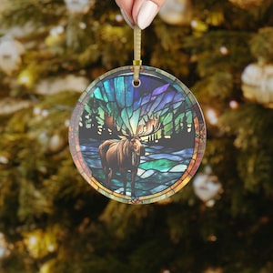 K&ouml;nnte beinhalten: Runder Weihnachtsbaumschmuck mit einem Buntglasdesign, das einen Elch zeigt, der in einer Waldszene mit einem blauen, gr&uuml;nen und violetten Farbschema steht.