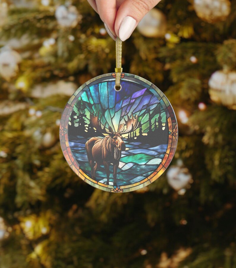 K&ouml;nnte beinhalten: Runder Weihnachtsbaumschmuck mit einem Buntglasdesign, das einen Elch zeigt, der in einer Waldszene mit einem blauen, gr&uuml;nen und violetten Farbschema steht.