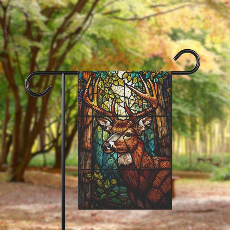 May include: A garden flag with a stained-glass effect depicting a deer with large antlers. The deer is brown and white, set against a backdrop of trees and foliage in green, blue, and brown. The flag is displayed on a black metal stand.