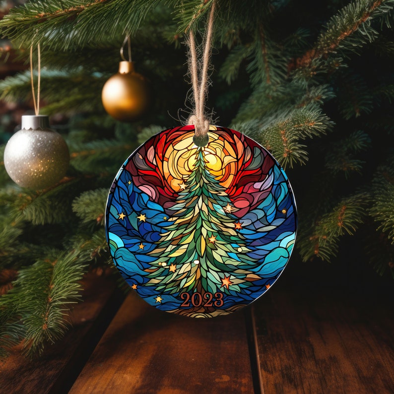 Puede incluir: Un adorno navide&ntilde;o redondo con un dise&ntilde;o de vitral de un &aacute;rbol de Navidad verde con una estrella amarilla en la parte superior. El &aacute;rbol est&aacute; rodeado de un fondo azul, rojo y amarillo con estrellas. El adorno tiene el a&ntilde;o 2023 escrito en &eacute;l.