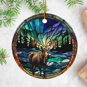 K&ouml;nnte beinhalten: Ein runder Keramik-Anh&auml;nger mit einem Buntglas-Design, das einen Elch zeigt, der in einem schneebedeckten Wald unter einem farbenfrohen Nordlicht steht. Der Anh&auml;nger ist perfekt, um Ihrer Weihnachtsdekoration einen Hauch von Natur zu verleihen.