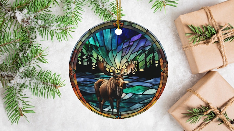 K&ouml;nnte beinhalten: Ein runder Keramik-Anh&auml;nger mit einem Buntglas-Design, das einen Elch zeigt, der in einem schneebedeckten Wald unter einem farbenfrohen Nordlicht steht. Der Anh&auml;nger ist perfekt, um Ihrer Weihnachtsdekoration einen Hauch von Natur zu verleihen.