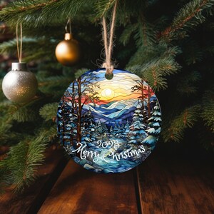 May include: Round Christmas ornament with a stained glass design of a winter forest scene. The ornament features a sunset over a mountain range with trees covered in snow. The text "2023 Merry Christmas" is written in white at the bottom of the ornament.