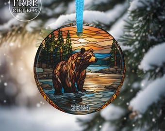 Könnte beinhalten: Ein runder Glas-Anhänger mit einem Buntglas-Motiv eines braunen Bären, der in einem See mit einem Sonnenuntergang im Hintergrund steht. Der Anhänger hat ein blaues Band und das Jahr 2024 ist auf der Unterseite aufgedruckt.