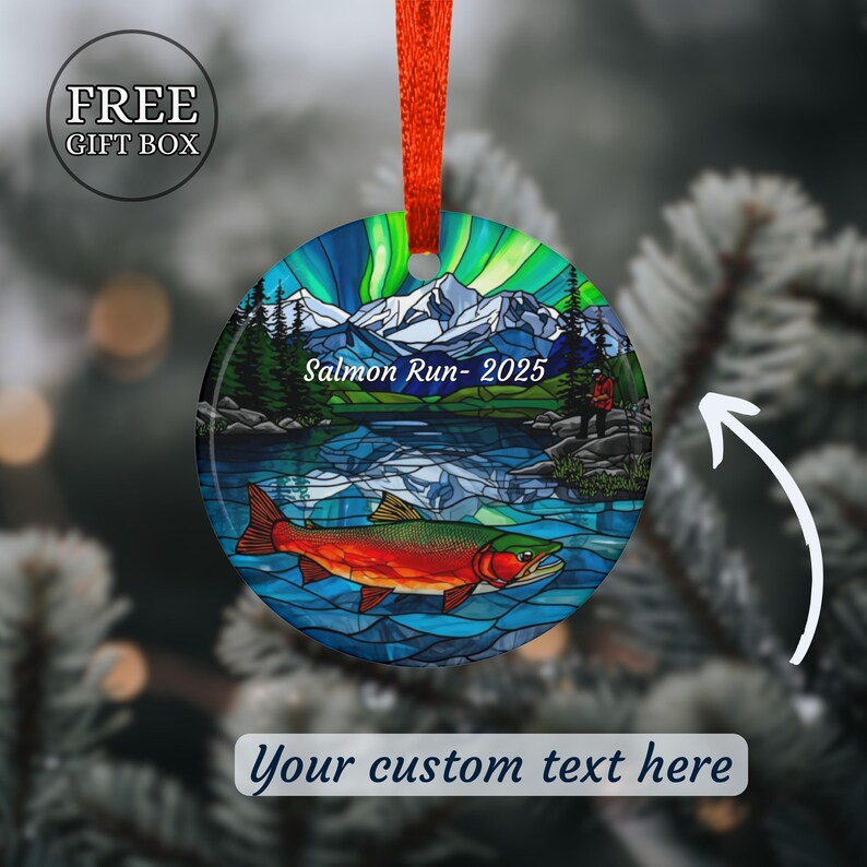 May include: Round Christmas ornament with a vibrant design. The ornament features a colorful illustration of a salmon in a lake, with mountains and the text "Salmon Run- 2025". It has a red ribbon for hanging and the words "Your custom text here".