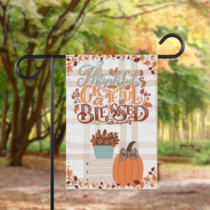 May include: A decorative garden flag with the words "Thankful, Grateful, Blessed" in a fall-themed design. The flag features a pumpkin with a bow, a flower pot, and autumn leaves. The flag is hanging on a black metal stand.