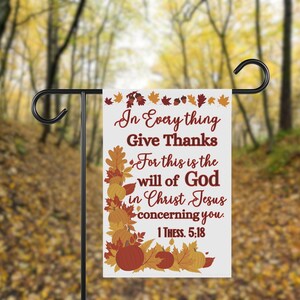 May include: A white garden flag with the text "In Everything Give Thanks" and a Bible verse. The flag features autumn leaves and pumpkins in shades of orange, brown, and yellow. The flag is displayed on a black metal stand.