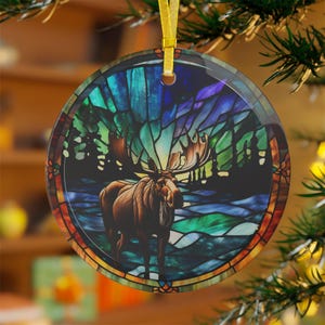 K&ouml;nnte beinhalten: Ein runder Glas-Anh&auml;nger mit einem Buntglas-Motiv eines Elchs, der in einem Wald mit einem farbenfrohen Nordlicht im Hintergrund steht.