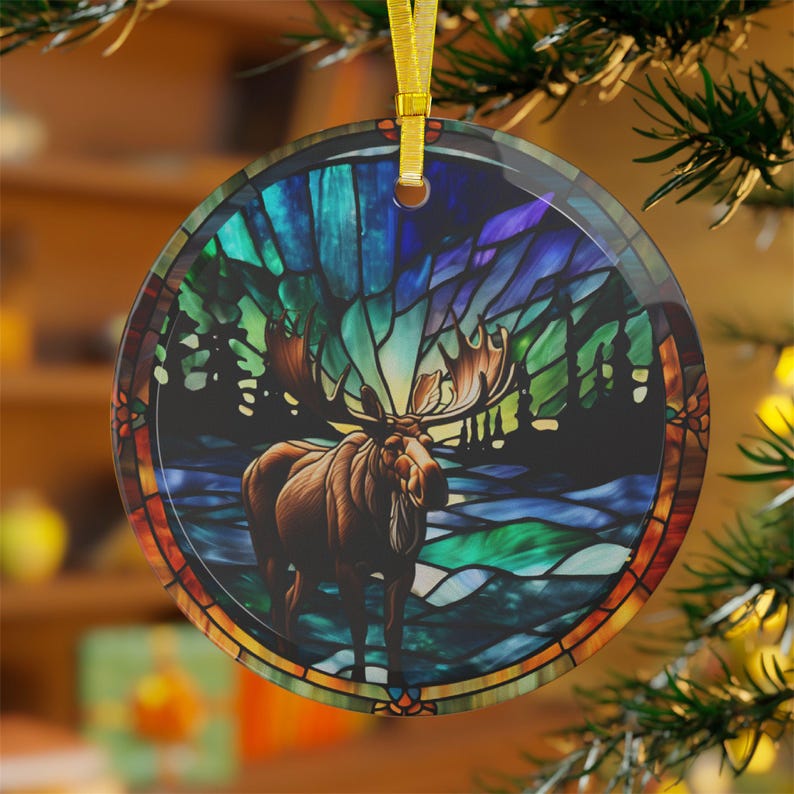 K&ouml;nnte beinhalten: Ein runder Glas-Anh&auml;nger mit einem Buntglas-Motiv eines Elchs, der in einem Wald mit einem farbenfrohen Nordlicht im Hintergrund steht.
