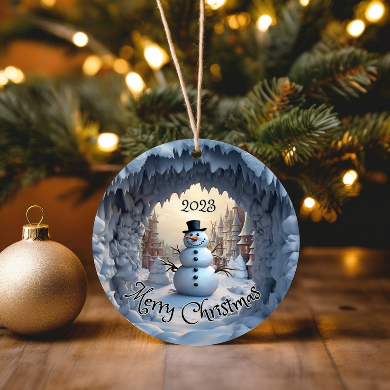 Puede incluir: Un adorno navide&ntilde;o redondo con un mu&ntilde;eco de nieve de pie en una escena nevada con una abertura en forma de cueva. El adorno tiene el texto "2023 Feliz Navidad" en &eacute;l.