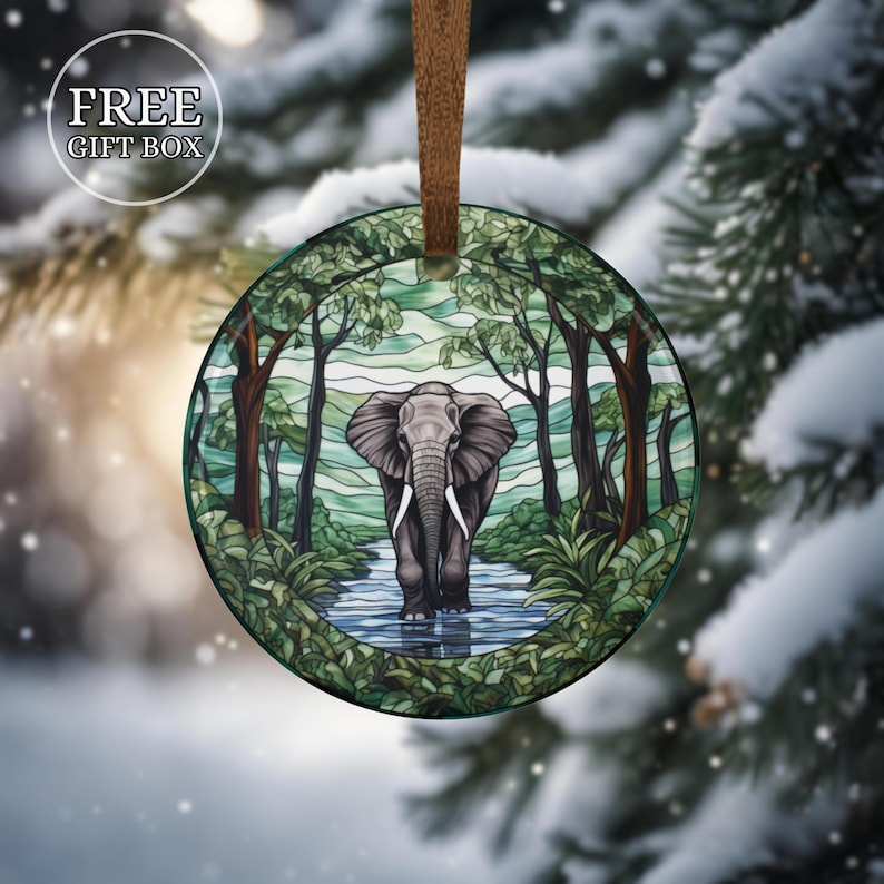 May include: Round glass ornament featuring an elephant walking in a river surrounded by trees. The ornament has a brown ribbon for hanging. The words "FREE GIFT BOX" are in a white circle in the upper left corner.