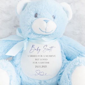 Può includere: Un orsacchiotto di peluche azzurro con un nastro abbinato. L'orso ha una toppa bianca sul petto con il testo "Baby Scott, portato per un momento, ma amato per sempre, 24.11.2023" in corsivo.