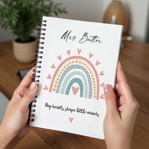 Può includere: Un quaderno a spirale bianco con il testo Mrs Butler in alto e Big hearts shape little minds in basso. Il design centrale mostra un arcobaleno multicolore nelle tonalità del rosa, giallo e verde acqua, circondato da piccoli cuori rosa.