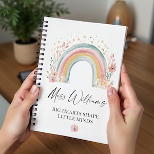 May include: Spiral-bound notebook featuring a watercolour rainbow design. The rainbow arches over the text "Miss Williams" and the quote "BIG HEARTS SHAPE LITTLE MINDS." Floral and star accents surround the rainbow. The notebook is held open.