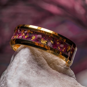 May include: A gold-toned ring featuring a central inlay of deep magenta crushed material interspersed with small, reflective gold-colored flakes. The band has a polished, metallic finish and a flat profile, showcasing a distinct contrast between the gold edges and the textured, colorful center inlay.