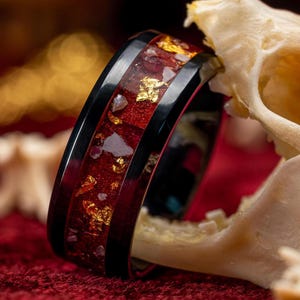 May include: A close-up of a black ring with a red and gold inlay. The ring features a black band with a red center filled with gold flakes and small, clear stones. The ring is set against a blurred background.