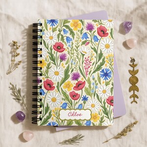 May include: A spiral-bound notebook with a floral design featuring red poppies, white daisies, and bluebells on a cream background. The name "Chloe" is written on a label. The notebook is surrounded by crystals and decorative elements.