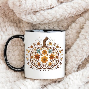 May include: White ceramic mug with a black handle and rim, featuring a floral pumpkin design in shades of brown, orange, and blue. The mug is set against a soft, textured background.