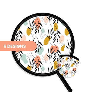 May include: A round white fabric item with a black border featuring a pattern of black leafy branches, mustard yellow, peach, and light blue abstract shapes, and small black dots. A smaller matching piece is attached, and text reads 6 DESIGNS.