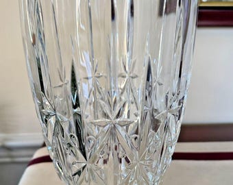 Puede incluir: Un jarrón de cristal transparente con un diseño de estrella. El jarrón tiene una abertura ancha y una forma ligeramente cónica. El diseño de estrella está grabado en el cristal, creando una superficie texturizada. El jarrón está sobre una superficie blanca.