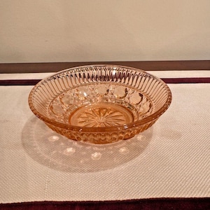 May include: A small, round, pink glass bowl with a decorative pattern. The bowl has a ribbed exterior and a starburst design in the center. The bowl is likely vintage or antique.