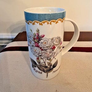 May include: Tall white ceramic mug with a floral design. The mug features a blue textured band at the top, a gold decorative trim, and a large handle. The floral design includes white and pink roses with the word "Rosa" and "Nouvelle" printed on the mug.