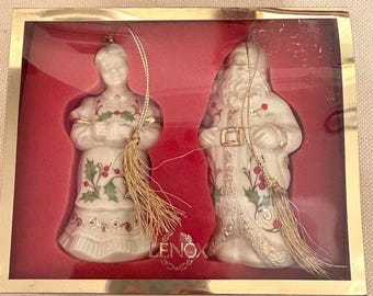 May include: A set of two white porcelain ornaments in a gold-trimmed box with a red interior. One ornament depicts a figure in a dress, the other a Santa figure, both featuring holly leaf and berry patterns, gold accents, and gold hanging tassels.