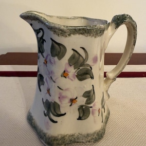 May include: A white ceramic pitcher with a floral design. The pitcher features a gray rim and handle, with hand-painted purple flowers and green leaves. The pitcher has a unique, slightly flared shape.