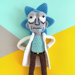 May include: A crocheted plush toy of a cartoon character with blue hair, a white lab coat, brown pants, and a light blue shirt. The toy has a droopy expression and large, white eyes.