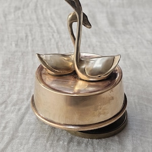 May include: A brass swan-shaped trinket dish. Two swans with intertwined necks form the handle, and their wings create the dish's bowls. The dish sits on a round, tiered base. The metal has a warm, golden hue.