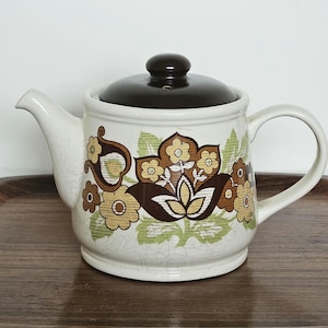 May include: Cream ceramic teapot with a dark brown lid and handle. The teapot features a floral pattern in brown, yellow, and green. The teapot is on a wooden tray.