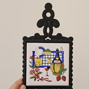 May include: Black metal trivet with a colourful tile insert. The tile features a still life with a wine bottle, cheese, strawberries, and a glass of red wine. Red flowers and a blue grid complete the design.