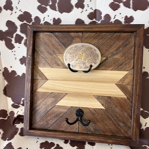 Op de afbeelding: Een vierkant houten wanddisplay met een donkerbruin gebeitste lijst en een geometrische inleg van lichte en donkere houten planken. Het bevat een zilverkleurige ovale riemgesp met gouden accenten en twee zwarte metalen haken.