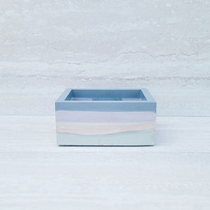 May include: A square, pastel-colored concrete box. The box is painted with horizontal bands of light blue, lavender, white, and mint green. The interior of the box is light blue. The box is sitting on a white surface.