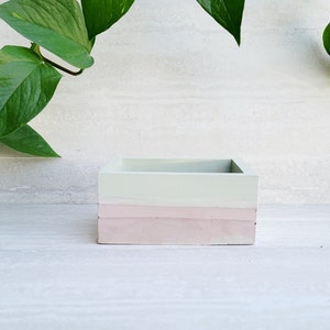May include: A square, three-toned planter with a light green top, a pink middle section, and a darker pink base. The planter is made of a solid material and is empty. Green leafy vines are in the background.