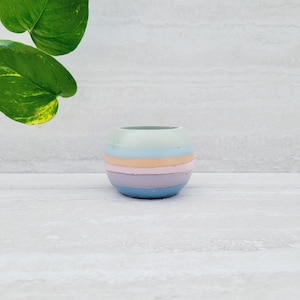 May include: A small, round, pastel-coloured planter with horizontal stripes of blue, lavender, peach, and mint green. The planter is made of concrete and sits on a white surface. Green leaves are visible in the background.