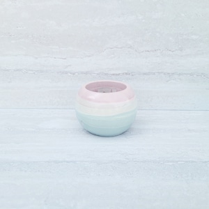 May include: A small, round ceramic candle holder in pastel shades. The holder is divided into three horizontal bands of colour: pink, white, and light blue. The interior is pink, and the holder sits on a light-coloured surface.
